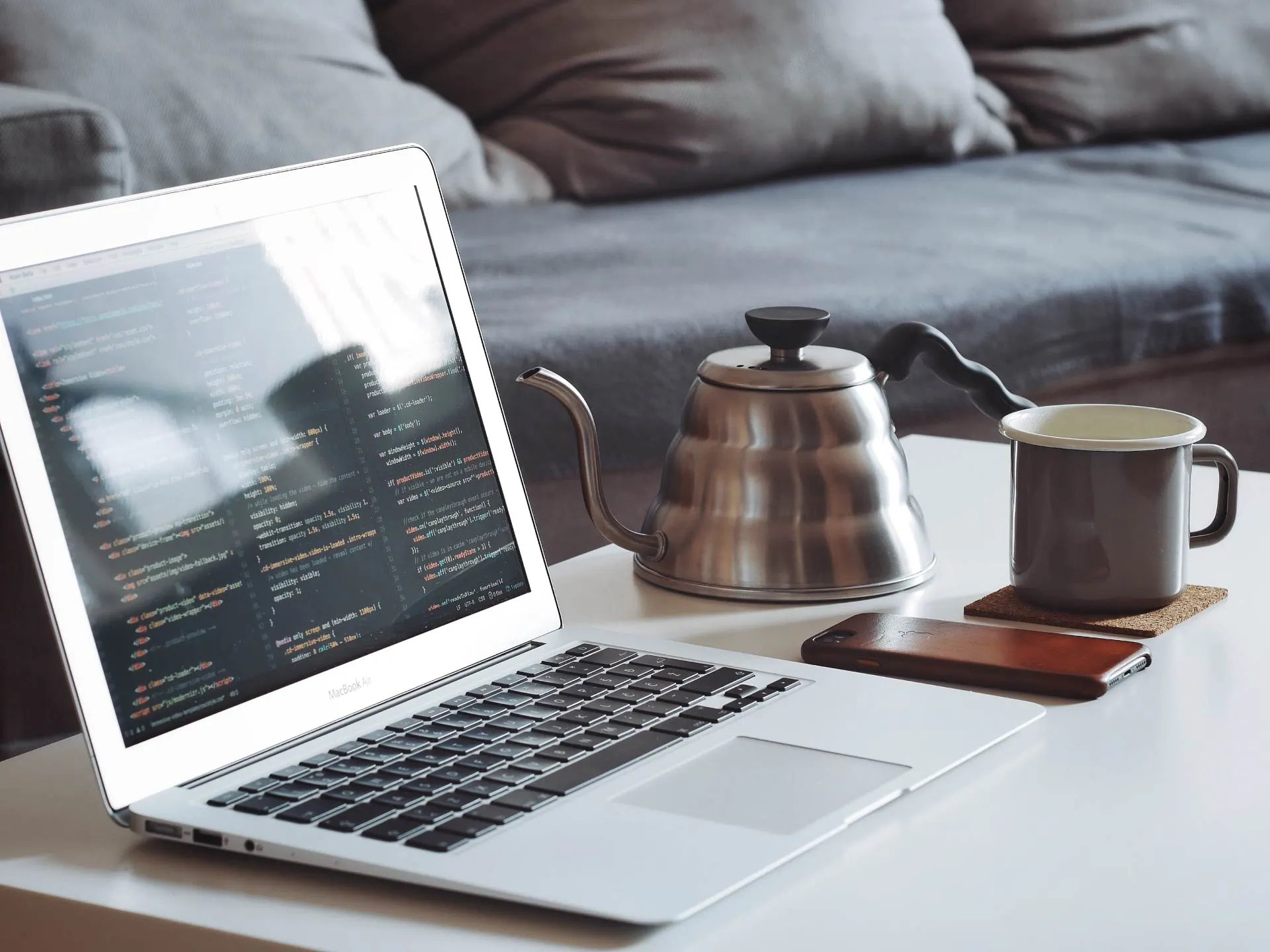 A laptop on a white table with a phone, silver teapot and mug to the right hand side in front of a sofa